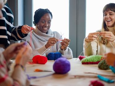 Happy multiracial group of friends enjoying craft workshop sitting at wooden table in cafe learning knitting with balls of wool, learning, togetherness, relaxation
