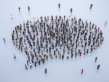 A large group of people dressed in formal and casual attire stand together, forming a unique shape on a smooth floor in a city setting, engaging in discussion or networking.