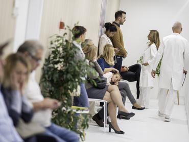 Egy orvosi rendelő várója páciensekkel; a háttérben egy doktornő beszélget egy fiatal férfival.