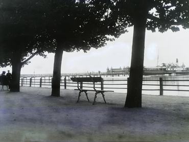 A kikötő és a fürdőház a mai Tagore sétányról nézve, Balatonfüred, 1911. Az előtérben pad, a tó vizén gőzhajó, a háttérben két sétáló alak.