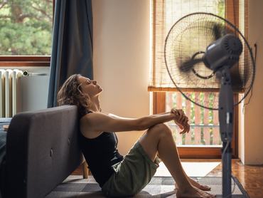 Egy hőgutától szenvedő fiatal nő otthon a padlón ül egy ventilátor előtt
