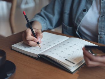 Rendezvényszervezői ütemterv és napirend 2024-re. Üzletasszony, aki mobiltelefonján nézi a naptárát, miközben az irodai asztalán lévő naptárba jegyzetel. Naptári eseményterv, munkatervezés