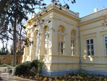 A A balatonfüredi Jókai Mór Emlékház kívülről. Az előtérben egy fa lombja, a ház világos színűre van festve.