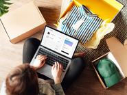 Felülnézet egy fiatal nőről, aki laptopján keresztül online fizetést hajt végre, miközben kicsomagolja a házhoz szállított csomagot. Online vásárlás és házhoz szállítás.